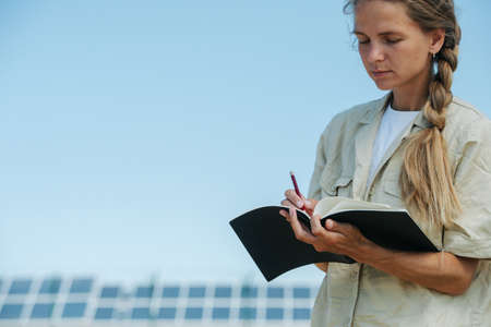 Young woman writing down things in her notepad. Over the sky view, low angle. Electric solar panels...