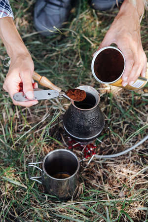 Hands adding coffee powder in a cezve with a metal folding spoon. On a grass while camping.