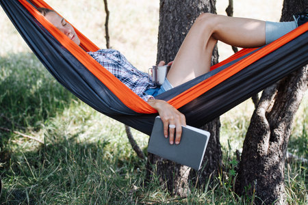 Dozed woman lying in a hammock, slugging in a shadow, between two big trees. She's stillholding a book and a cup in her hands.の写真素材