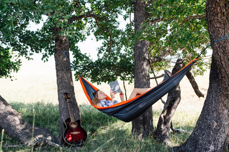 Napping woman lying in a hammock, slugging in a shadow, between two big trees. She's holding book in her hands, eyes closed.の写真素材