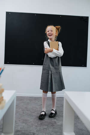 Exhilarated schoolgirl hugging textbook standing in front of a balckboard in a classroom.の写真素材
