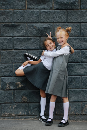 Portrait of two cute little schoolgirl friends. One is lifting her leg behind, the other is giving her horns. Posing in front of the painted brick wall.の写真素材