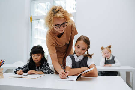 Teacher helping a schoolgirl sitting behind a desk, writing in her notebook. Frontal view.の写真素材