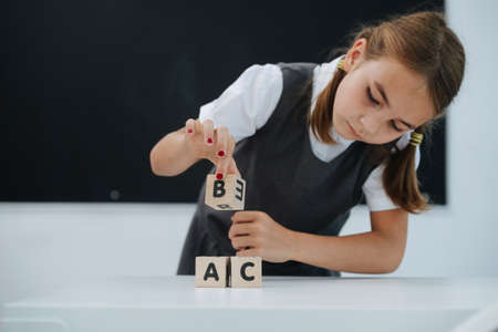 Focused little shoolgirl playing on a desk with toy letter cubes. Frontal view. Blackboard in background.の写真素材