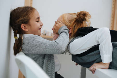 Active elementary schoolgirls playing with each other. One is getting in a face, second one is covering eyes with palms. Side view.の写真素材