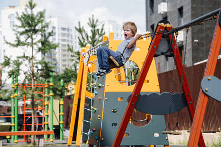 Yelling little boy riding on swings on a children playground. He is flying high, bending chain...