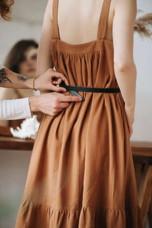 Body image of a woman in brown dress, and hands helping to tighten her waist belt. No head. Blurred face reflection in the mirror.の写真素材