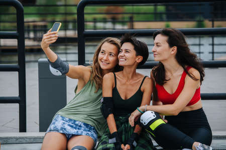 Three smiling skater girls taking selfie, leaning on the railing at skatepark .. Sitting close to each other, looking at the phone camera.の写真素材