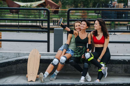 Skater girls taking selfie, leaning on the railing at skatepark. Sitting close to each other.の写真素材