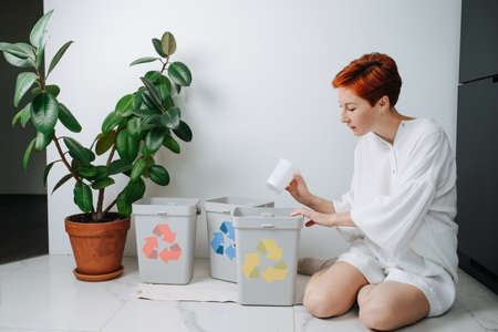 Concerned woman sitting on her legs, sorting garbage beween small recycle bins at home. They have differently colored arrows on them. Holding plastic cup.の写真素材