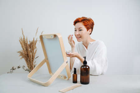 Lively short-haired woman looking at the mirror with a wooden frame, applying the concealer. Using eco-friendly products and things.の写真素材