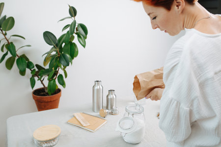 Woman pouring white powder in a jar from a paper bag. Eco friendly objects on the table.の写真素材