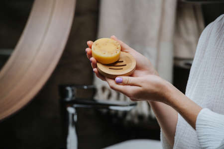 Round soap dish in hands of a mature woman with an eco soap block. She's standing in front of the...