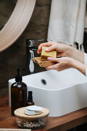 Wooden soap dish in hands of a mature woman with an eco soap block. She's standing in front of the sink in a bathroom.の写真素材