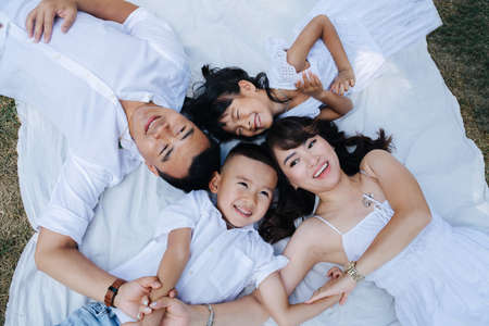 Top view portrait of a joyous asian family in white clothes lying on a blanket in a park, enjoying last warm days of the early fall.の写真素材