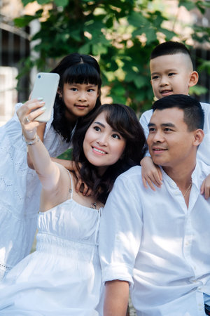 Beautiful asian family in white clothes taking selfie in a park. They are having picnic, enjoying...