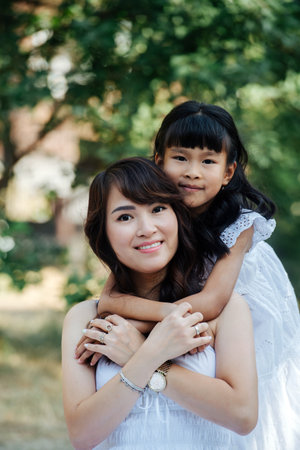 Cute portrait of a little girl hugging her mother from behind. They both wearing white clothes having picnic in a park, enjoying last warm days of the early fall in tree shadow.の写真素材