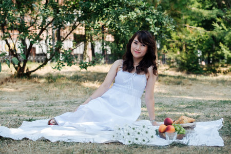 Elegant portrait of a uplifted lovely asian woman sitting on a blanket in a park. She's wearing white dress, having picnic in a park, enjoying last warm days of the early fall in tree shadow.の写真素材