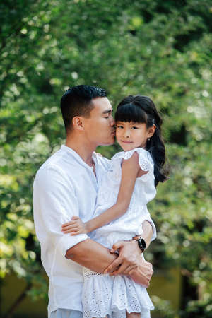 Loving asian father holding his daughter in his hands, kissing her in a cheek. She's looking at the camera. They are spending time in a park, enjoying last warm days of the early fall.の写真素材