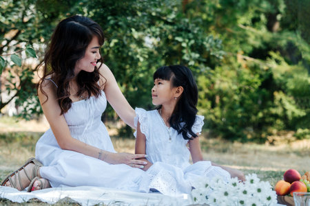 Asian mom and daugther in white clothes having picnic in a park, enjoying last warm days of the early fall in a tree shadow.の写真素材