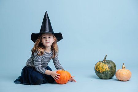 Cute little girl in witch hat playing with pumpkins, sitting on the floor. She's dressed for halloween. Over blue background.の写真素材