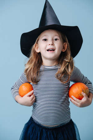Little girl in a witch hat holding two small pumpkins, she's dressed for halloween. Over blue background.の写真素材