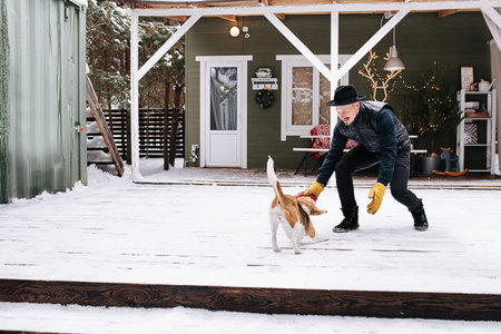 Friendly man in a hat and leather jacket playing with a happy beagle dog. In front of the house on a snowy plank floor.の写真素材