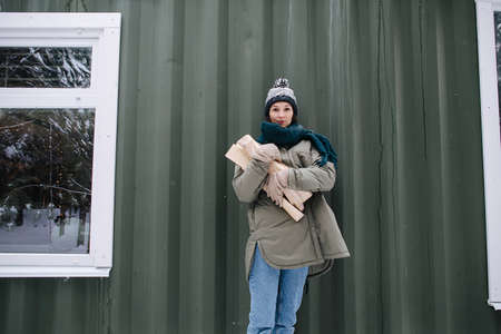Longshot portrait of a woman in snowy winter clothes holding logs in hands. She wears knitted hat and scarf. Standing in front of a house wall.の写真素材