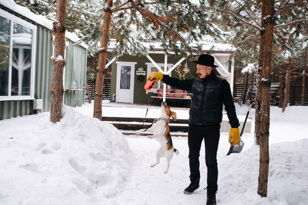 Dressed in black man playing with a happy beagle dog, teasing her, making jump. In front of the house on a snowy path between treesの写真素材