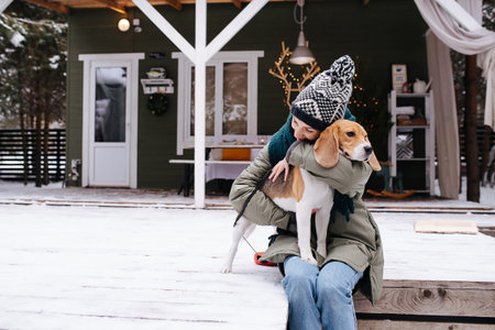 Gentle woman sitting on a snowy terrace in front of the house, hugging her beagle dog standing on her lap, leaning to her.の写真素材