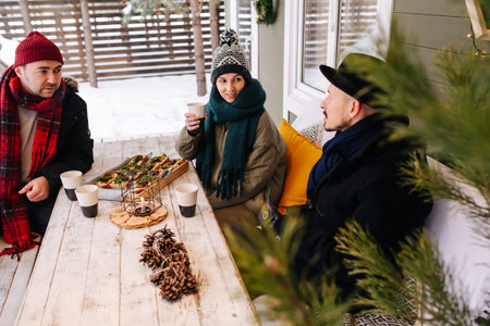 Good middle aged friends sitting on a snowy house terrace under a roof, talking to each other, drinking coffee. Trees in background.の写真素材
