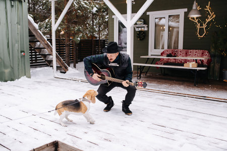 Eccentric musician in a hat and leather jacket playing guitar for a friendly dog. He is standing in front of the house on a snowy plank floor.の写真素材