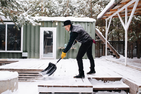 Man in a hat and leather jacket clearing snowy plank floor in front of house with a plastic wide shovel.の写真素材