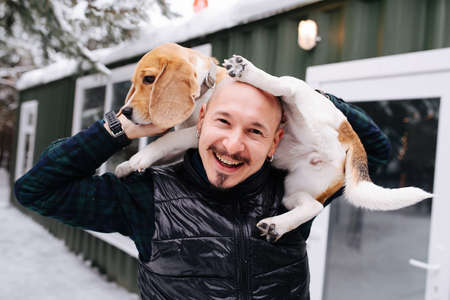 Cheerful bold man playing with a dog, lifting her on his shoulders, she's trying to hold balance or get out. Outside a house at winter.の写真素材