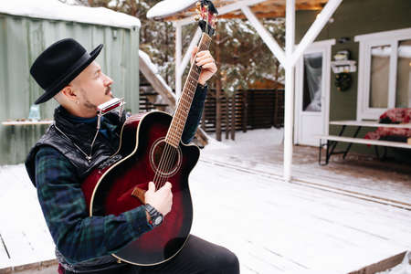 Cool looking musician in a hat and leather jacket playing guitar in front of the house on a snowy plank floor. Angling his guitar up.の写真素材