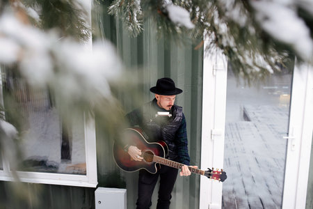 Reflective musician in a hat and leather jacket playing guitar next to a house door on a snowy plank floor. Holding guitar.の写真素材