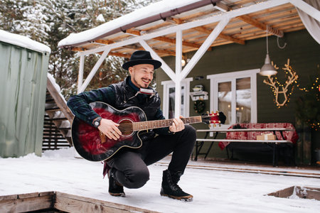 Radiant musician in a hat and leather jacket crouching with guitar in front of the house on a snowy plank floor. Smiling, playing for someone.の写真素材