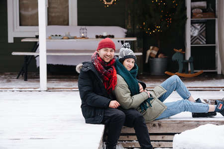 Happy comfortable couple sitting on a snowy terrace step in front of the house, posing for a photo. Both wearing hats, jackets and scarfs. Woman leaning with her back on the man.の写真素材