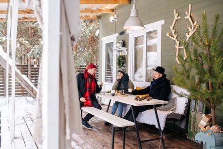 Friends sitting on a snowy house terrace under a roof, talking to each other, drinking coffee. Trees in background.の写真素材
