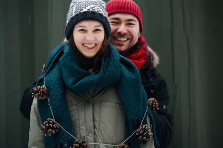 Portrait of a funny middle age couple standing outdoors at winter. Woman is wearing a cone necklace. Both wearing hats, jackets and scarfs. In front of corrugated sheet house wall.の写真素材