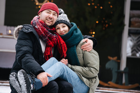Thirty year old couple sitting on a snowy terrace step in front of the house, posing for a photo. Both wearing hats, jackets and scarfs. He's holding her ankles on his knees.の写真素材