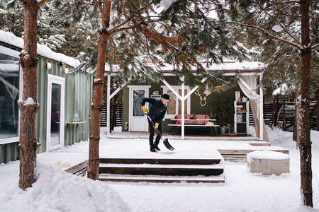 Longshot of a man in a hat and leather jacket clearing snowy plank floor in front of house with a plastic wide shovel.の写真素材
