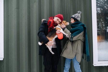 Funny couple trying to pose for a photo with their beagle outdoors at winter. Man is hugging the dog, woman is calming her down. In front of corrugated sheet house.の写真素材