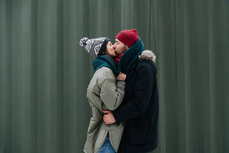 Passionate couple in love kissing outdoors at winter. Both wearing hats, jackets and scarfs. In front of corrugated sheet house wall.の写真素材