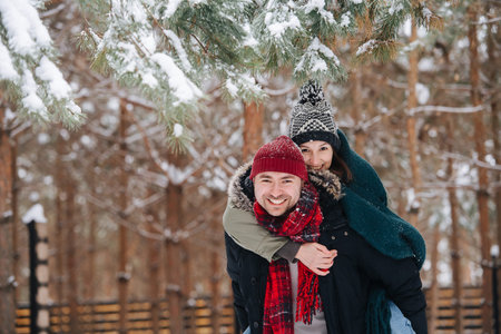 Cheeky guy lends his back for the woman to ride. Both smiling. In a conifer tree park at winter. Both wearing hats, jackets and scarfs. Below snowy branches.の写真素材