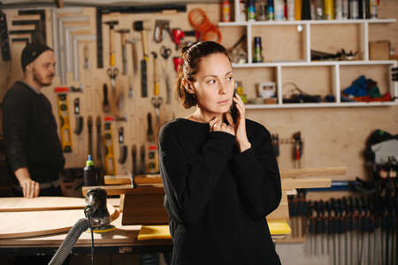Woman client speaking on the phone while being in a carpentry workshop. Crew in background. Tools hanging on the wall.の写真素材