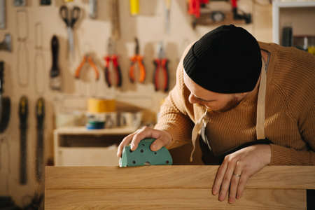 Attentive carpenter in a watch cap grinding a piece of wood, blurred background. In a big workshop. Number of tools hanging on the wall.の写真素材