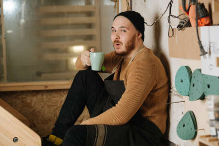 Funny carpenter in a watch cap sitting on a wooden stairs in a big workshop, drinking tea from the mug. Looking at the camera with a funny face expression. Number of tools hanging on the wall.の写真素材