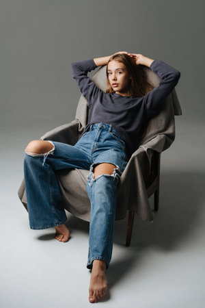 Chill teenage girl sitting on an armchair, holding hands over her head. Portrait, looking at the camera. Wearing mom jeans with holes on her knees and grey long-sleeve.の写真素材