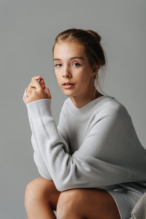 Subtly smiling teenage girl sitting on a chair, leaning forward, looking at the camera. She's wearing warm longsleeve covering her body down to her thighs with bare legs showing under.の写真素材